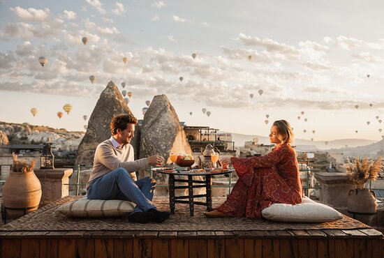 A young couple sits with their backs turned to the camera by the entrance of a cave hotel in Cappadocia, Turkey, admiring the unique rock-carved architecture in a peaceful and historic setting.